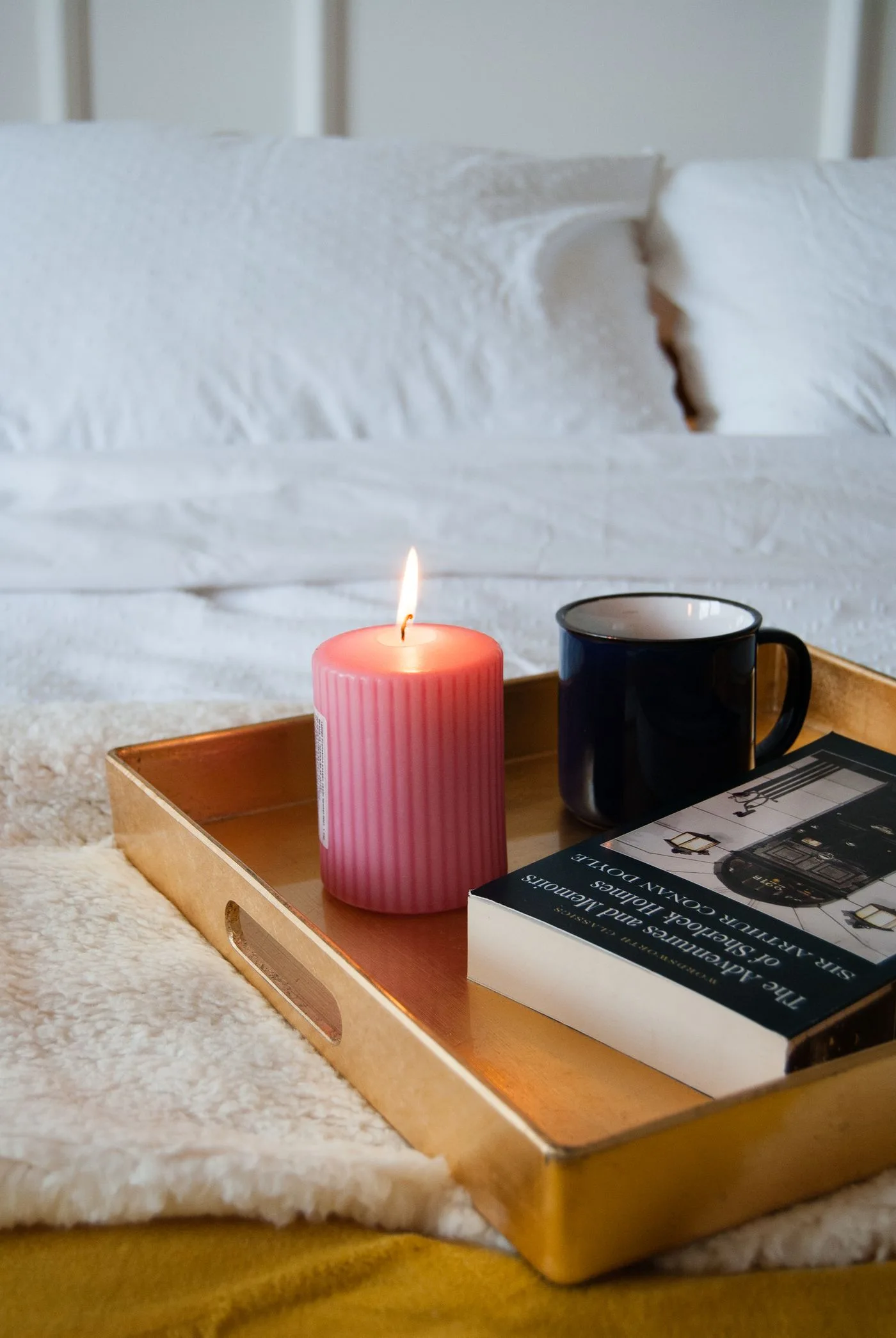 A book, a mug and a pink candle on a tray over pale bedding